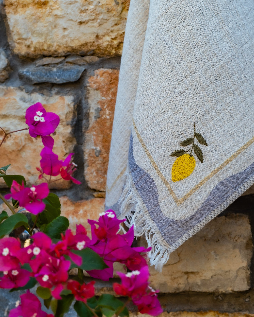 Kitchen towel with a hand stitched lemon decal against a stone wall with flowers in the foreground