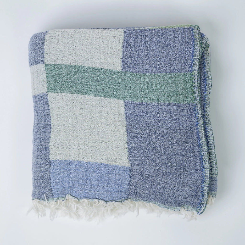 A cotton throw blanket with a block pattern in teal, gray, and blue colors, featuring a fringed edge.