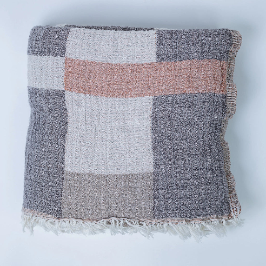 A cotton throw blanket with a block pattern in beige, gray, and coral colors, featuring a fringed edge.