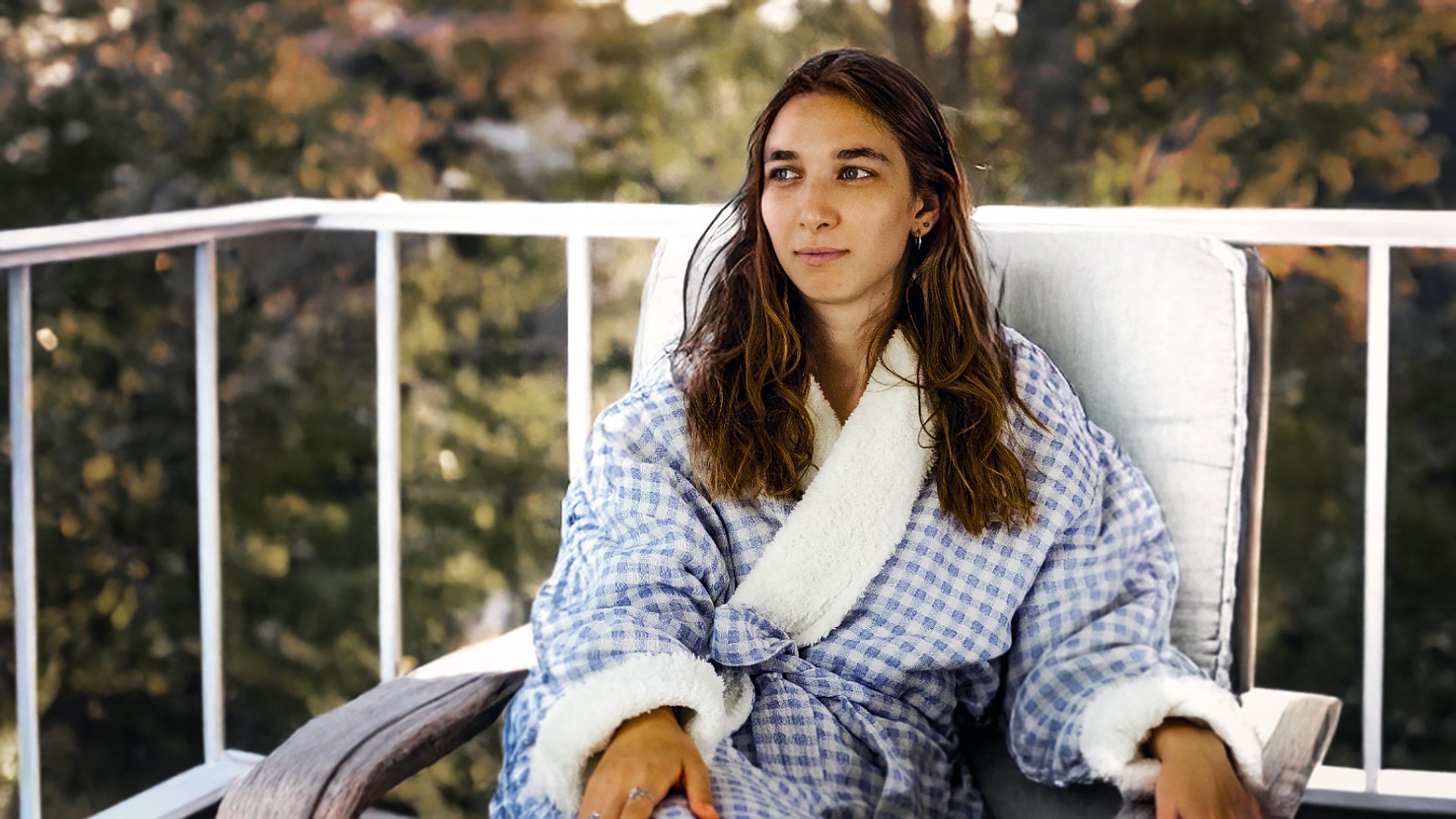 Woman in a checkered robe sitting on a chair outdoors with trees in the background