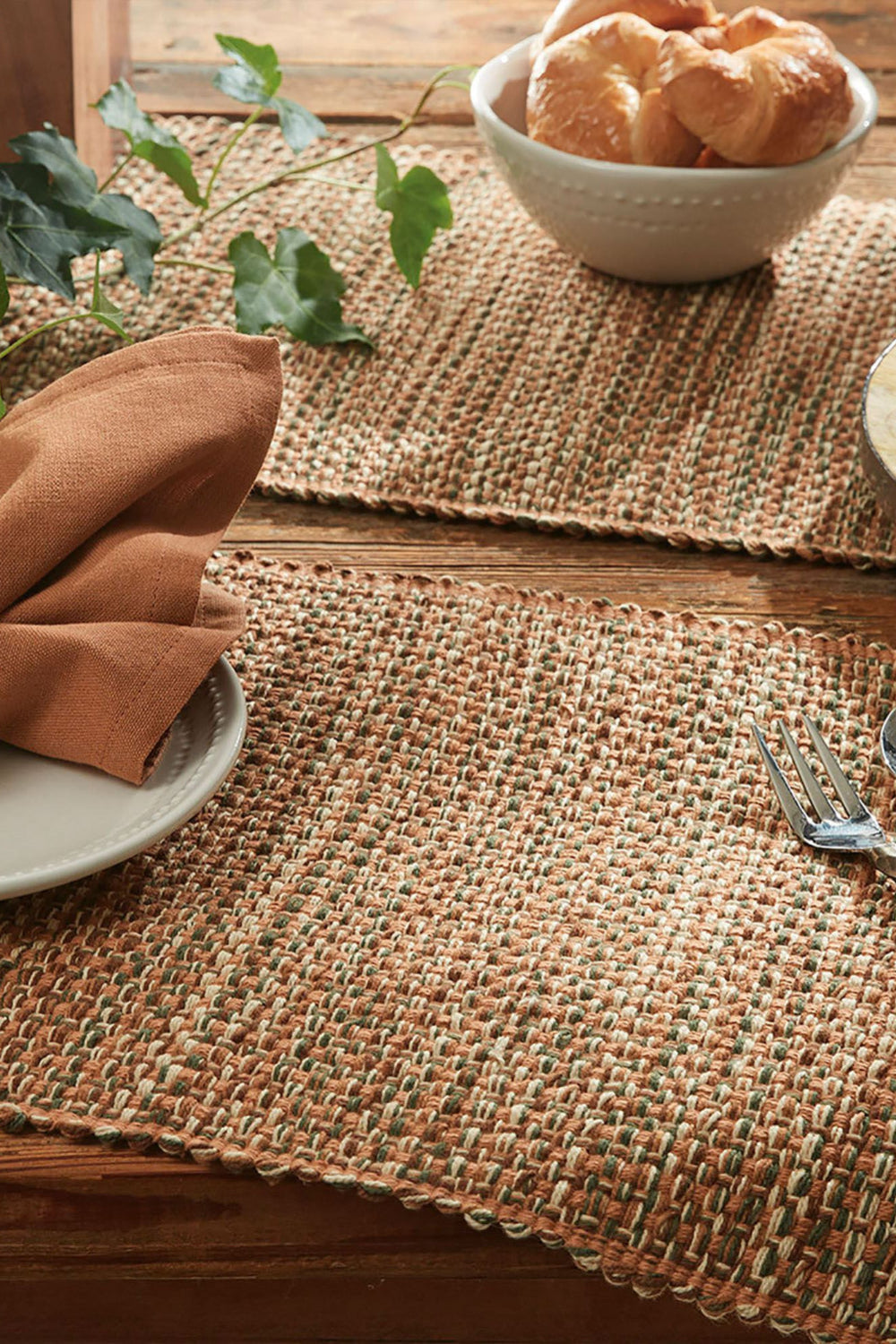 Table setting with woven placemats, croissants, grapes, and cutlery on a wooden table.