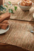 Table setting with woven placemats, croissants, grapes, and cutlery on a wooden table.
