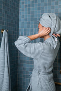 Person in a blue waffle bathrobe with a towel on their head in a bathroom with blue tiles.