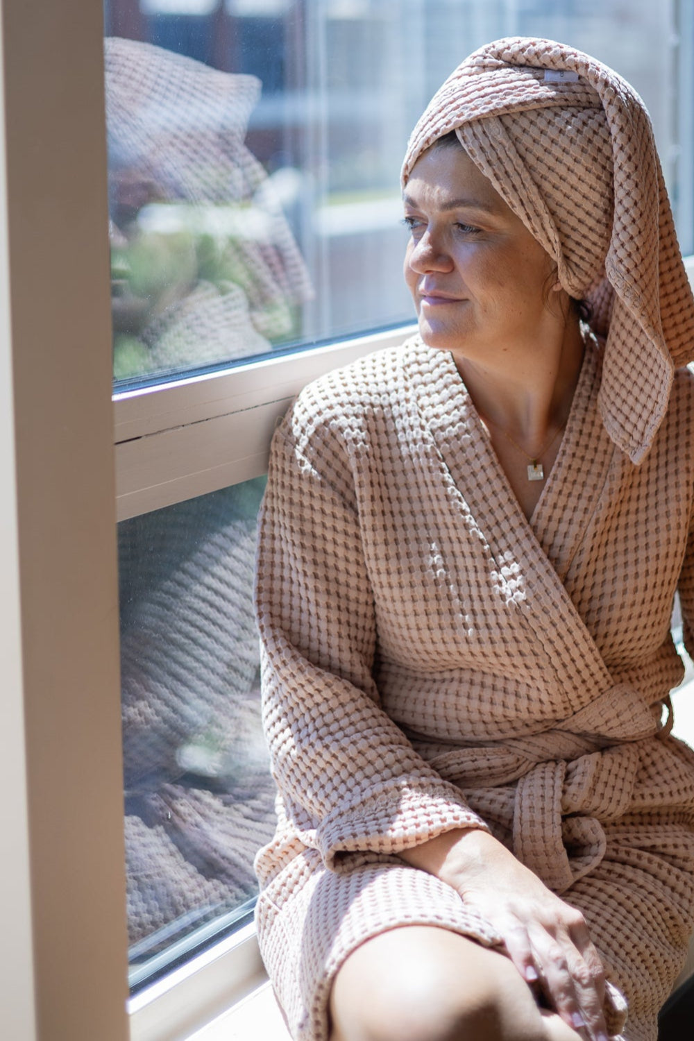 Woman in a checkered robe sitting by a window, looking out.